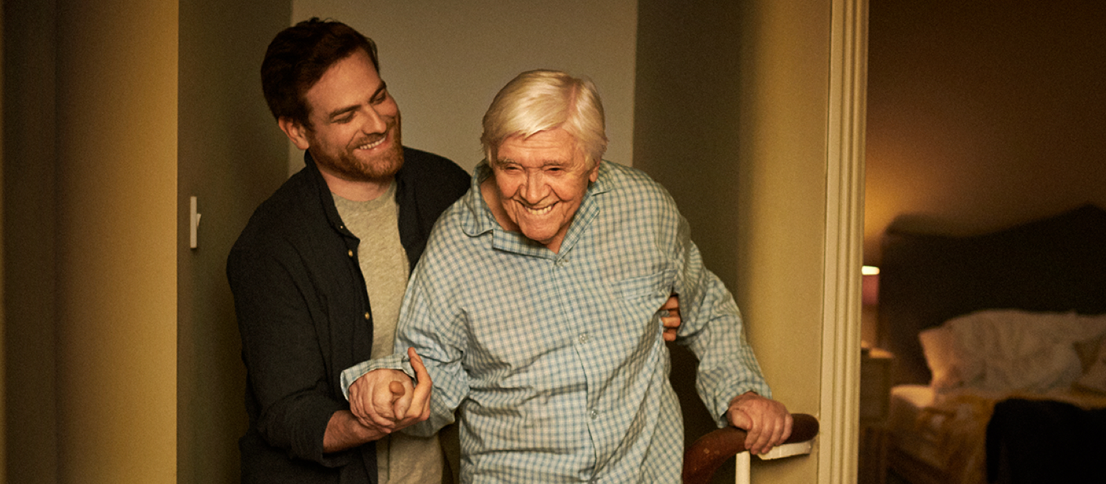A smiling young man helps an older man in pyjamas upstairs toward a bedroom. A smiling young man helps an older man in pyjamas upstairs toward a bedroom.